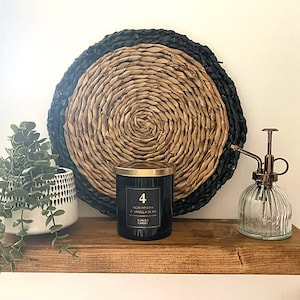 May include: A decorative shelf displays a black candle with gold lid, a glass mister, and a potted plant. A woven wall hanging with black and natural tones is behind the candle. The candle label reads "Noir Myrrh & Vanilla Bean".