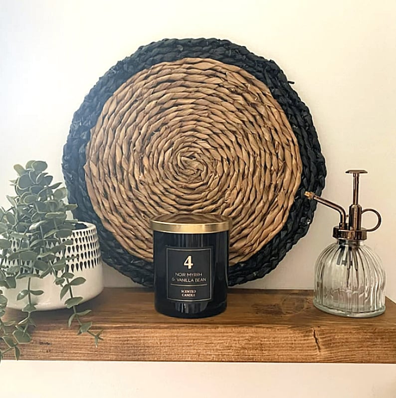 May include: A decorative shelf displays a black candle with gold lid, a glass mister, and a potted plant. A woven wall hanging with black and natural tones is behind the candle. The candle label reads "Noir Myrrh & Vanilla Bean".