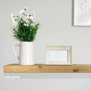 May include: A wooden shelf displays a white pitcher with daisies, a framed photo, and the text "old pine." The shelf is made of light-colored wood, creating a minimalist aesthetic. The frame has a distressed, textured finish.