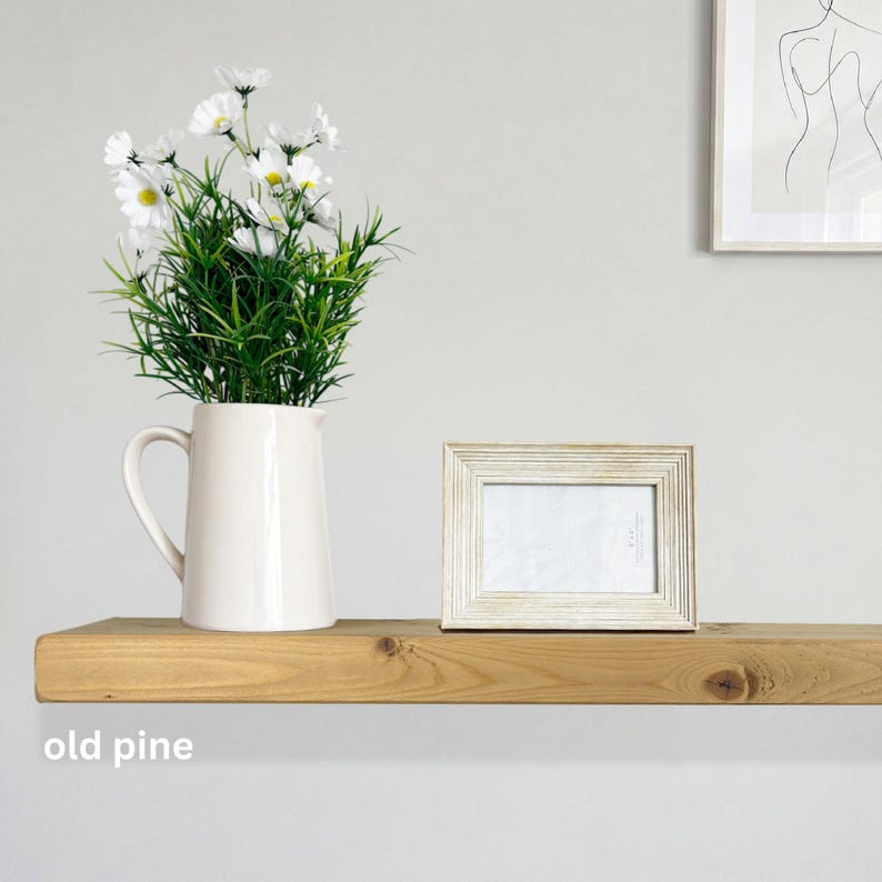 May include: A wooden shelf displays a white pitcher with daisies, a framed photo, and the text "old pine." The shelf is made of light-colored wood, creating a minimalist aesthetic. The frame has a distressed, textured finish.