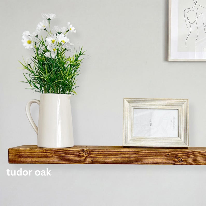 May include: A wooden shelf with a cream-colored pitcher holding white flowers and green foliage. A framed photo sits to the right. The shelf is labeled "tudor oak".