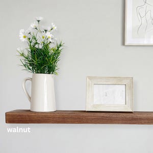 May include: A wooden shelf with a dark walnut finish, holding a white pitcher with white flowers and a white framed photo. The word "walnut" is printed on the shelf.