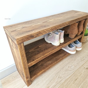 May include: Dark oak wood shoe rack with two shelves. The shoe rack has a rustic finish and is holding a pair of pink trainers, a pair of brown boots, and a pair of white trainers.