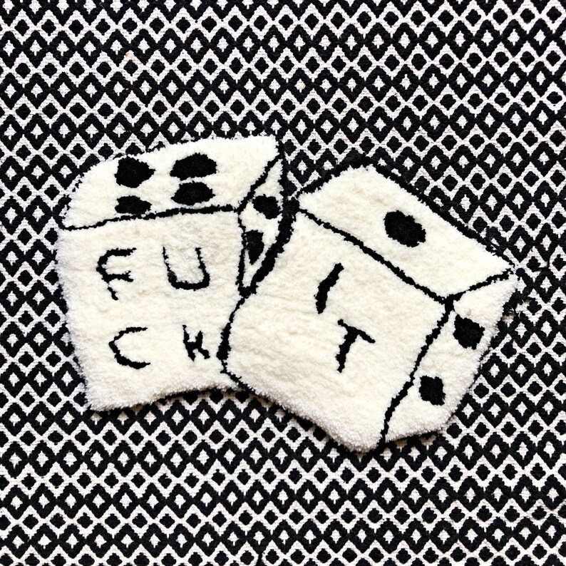 May include: Two white dice with black dots and the words "F.U.C.K.I.T." embroidered on them. The dice are on a black and white patterned background.