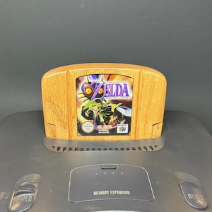 May include: A wooden Nintendo 64 game cartridge featuring "The Legend of Zelda: Majora's Mask" game label. The cartridge is set on a gray Nintendo 64 console, with the "Power" and "Reset" buttons visible.