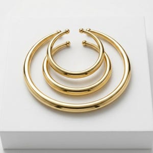 Puede incluir: Tres pulseras doradas abiertas de diferentes tamaños están dispuestas sobre una superficie blanca. Las pulseras tienen un acabado liso y pulido y extremos redondeados. La pulsera más grande está en la parte inferior, con las otras dos anidadas encima.