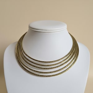 Könnte beinhalten: Mehrreihige Halskette mit strukturierter, antikgoldener Oberfläche. Die Halskette wird auf einer weißen Büste präsentiert, die ihr mehrschichtiges Design und die komplizierten Details hervorhebt. Die Halskette hat einen klassischen, eleganten Stil.