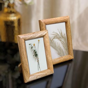 May include: Two wooden picture frames with light brown wood grain. One frame displays a sprig of dried flowers, the other a palm frond. The frames are on a dark surface, with a gold vase in the background.