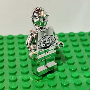 May include: A shiny silver Lego figure of C-3PO from Star Wars. The figure has a detailed design, including a circular pattern on the chest and round silver eyes. It stands on a green Lego baseplate.