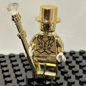 Könnte beinhalten: Eine goldfarbene Lego-Figur mit Zylinder und einem Gehstock mit einem klaren Diamanten an der Spitze. Die Figur trägt eine Weste mit schwarzen Details und weiße Handschuhe. Sie steht auf einer schwarzen Lego-Basis.