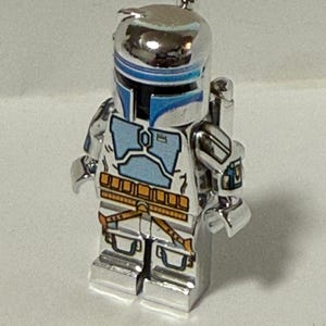 May include: A silver action figure of a character with a helmet and armour. The figure has blue and white accents on the helmet and chest, with brown and yellow details on the belt and legs. A collectible toy.