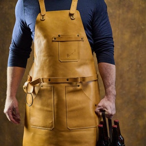 May include: A tan leather apron with adjustable straps and multiple pockets. The apron has a waist belt with a metal ring and a small pocket with a logo. The apron is worn over a blue long-sleeve shirt.