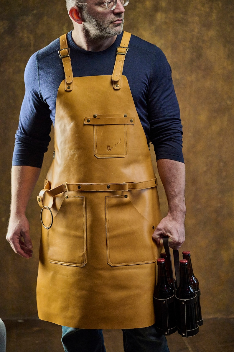 May include: A tan leather apron with adjustable straps and multiple pockets. The apron has a waist belt with a metal ring and a small pocket with a logo. The apron is worn over a blue long-sleeve shirt.