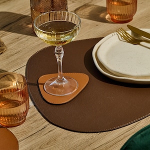 May include: A dining table setting with a champagne glass on a brown leather placemat, a stack of white plates, and gold cutlery. A small orange glass and a wooden bowl with blueberries are also present.