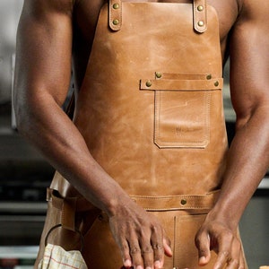 May include: A person wearing a brown leather apron with a large pocket and brass buckles is seasoning a raw steak with peppercorns on a wooden cutting board.