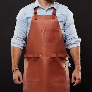 May include: A brown leather apron with two front pockets and a chest pocket. The apron has adjustable straps and a waist belt. The word "Margot" is written on the chest pocket. The apron is worn over a light blue denim shirt.