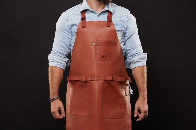 May include: A brown leather apron with two front pockets and a chest pocket. The apron has adjustable straps and a waist belt. The word "Margot" is written on the chest pocket. The apron is worn over a light blue denim shirt.