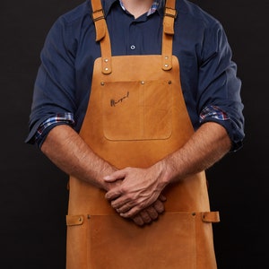 May include: A tan leather apron with adjustable straps and two front pockets. The apron has a large front pocket and a smaller pocket. The word "Margal" is handwritten on the front pocket. The apron is worn over a blue shirt.