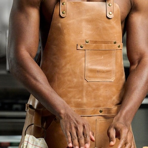 May include: A tan leather apron with adjustable straps and brass hardware. The apron features a front pocket and is worn while preparing raw meat. The image shows a person preparing food.