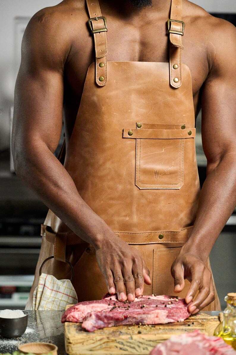 May include: A tan leather apron with adjustable straps and brass hardware. The apron features a front pocket and is worn while preparing raw meat. The image shows a person preparing food.