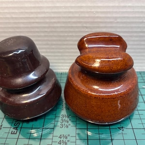 May include: Two antique brown ceramic insulators. One is a dark, glossy brown, and the other is a lighter, more reddish-brown. They are sitting on a green and white measuring surface.
