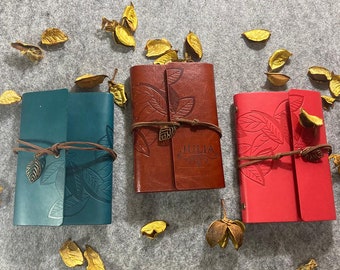May include: Three leather journals in teal, brown, and red, each with a leaf design and a leather tie closure. The brown journal has the name "JULIA" embossed on it. Dried leaves are scattered around the journals.