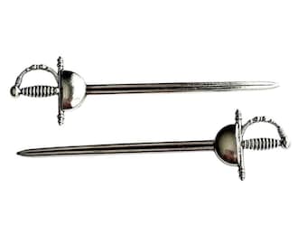 May include: Two silver-colored sword-shaped letter openers. Each opener has a detailed hilt and a long, pointed blade. The design is reminiscent of a historical rapier.