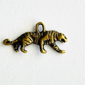 May include: A gold-toned metal charm in the shape of a tiger. The tiger is walking with its head turned to the side. The charm has a loop at the top for attaching to a necklace or bracelet.