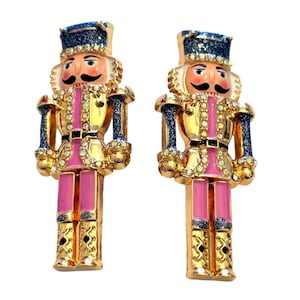 May include: A pair of nutcracker-shaped earrings featuring gold-tone metal, pink enamel accents, and blue glitter detailing on the hats and sleeves. The figures are embellished with clear rhinestones along the jackets and hats, with painted faces.