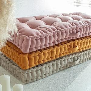 Op de afbeelding: Een stapel van drie vierkante vloerkussens in roze, goud en grijs. Elk kussen heeft een gecapitonneerd ontwerp en een stoffen handvat. De kussens zijn op elkaar gestapeld.