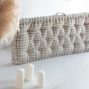 May include: A rectangular, tufted cushion with a gray and white gingham pattern. The cushion has a handle and is accented with a ruffled edge. Three white pillar candles are in the foreground.