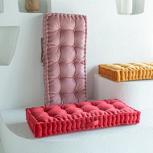 May include: A collection of tufted cushions in various colors. The image features a long, rectangular dusty rose cushion, a rectangular red cushion, and a smaller, square gold cushion. The cushions have a button-tufted design and are made of a soft, plush material.
