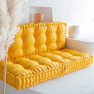 Puede incluir: Un cojín de suelo amarillo acolchado con un asiento y respaldo gruesos y acolchados. El cojín está hecho de una tela suave y cómoda y tiene un diseño simple y moderno.