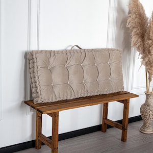 May include: A rectangular beige tufted cushion with button detailing and a handle, resting on a wooden bench. The cushion has a scalloped edge. The bench is made of dark wood. Decorative pampas grass is in a vase in the background.