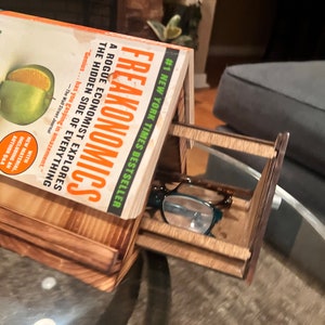 May include: A wooden house-shaped glasses holder with a book titled "Freakonomics" by Steven D. Levitt and Stephen J. Dubner resting on top. A pair of eyeglasses is inside the holder.
