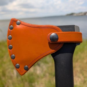 May include: A tan leather axe sheath covering the blade of a black axe. The sheath features a curved edge secured with seven dark metal rivets along the perimeter and a single matching metal snap button closure on the strap wrapping around the handle.