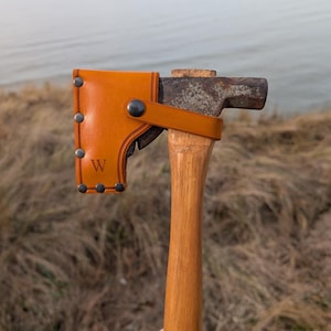 May include: A metal hatchet with a wooden handle featuring a tan leather sheath covering the blade. The sheath is secured with dark metal rivets and a snap button, and it is embossed with the letter W on the lower left side.