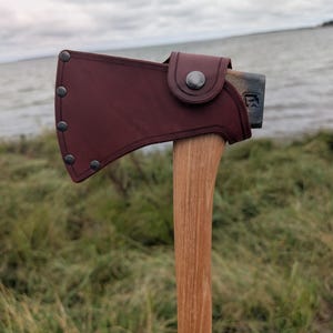 May include: A hand axe with a light-colored wooden handle and a dark maroon leather sheath covering the metal head. The sheath is secured with five metal rivets along the curved edge and a single metal snap button closure on the side.