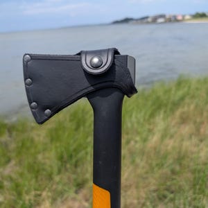 May include: A black hatchet with a protective black leather sheath covering the blade. The sheath is secured with metal rivets and a snap button closure. The handle of the hatchet is black with a bright orange section visible at the bottom.