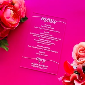 May include: A clear acrylic menu card with white script. The menu lists starters, entrées, accompaniments, and dessert options. The card is set against a bright pink background and surrounded by pink and red flowers. The menu is for Jazmin & Adolfo, October 29, 2022.
