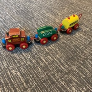 Puede incluir: Un tren de juguete de madera vintage con tres vagones. El vagón motor es marrón con una chimenea negra y las palabras "MAGIC" impresas en el lateral. El segundo vagón es verde con la palabra "COAL" impresa en el lateral. El tercer vagón es amarillo con un tanque negro y una manija negra.