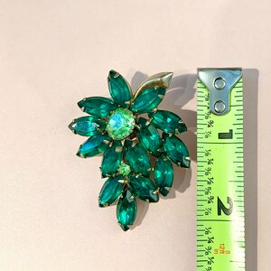 Pode incluir: Um broche em tom dourado e verde em forma de cacho de flores. O broche mede aproximadamente 5 cm de comprimento.