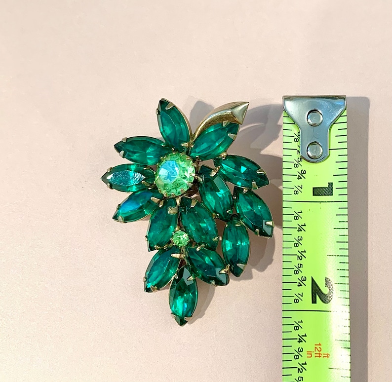 Pode incluir: Um broche em tom dourado e verde em forma de cacho de flores. O broche mede aproximadamente 5 cm de comprimento.