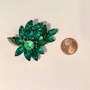 Pode incluir: Um broche de strass verde em forma de flor com uma configura&ccedil;&atilde;o em tom dourado. O broche &eacute; aproximadamente do tamanho de uma moeda de um centavo de d&oacute;lar americano.