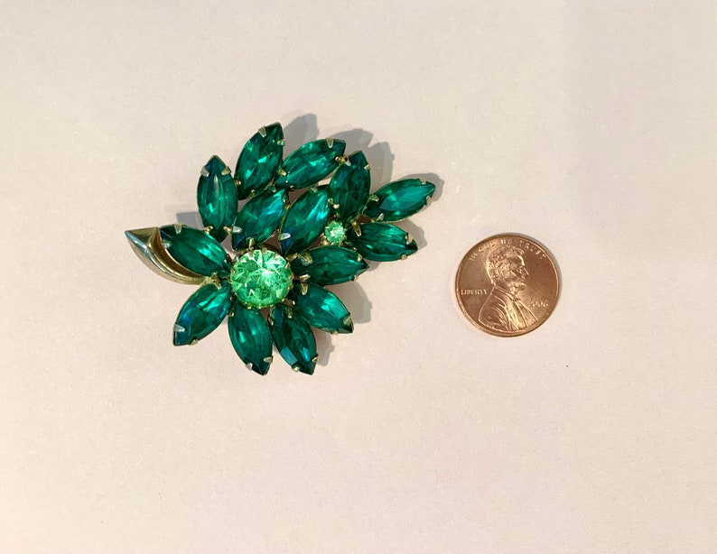 Pode incluir: Um broche de strass verde em forma de flor com uma configura&ccedil;&atilde;o em tom dourado. O broche &eacute; aproximadamente do tamanho de uma moeda de um centavo de d&oacute;lar americano.