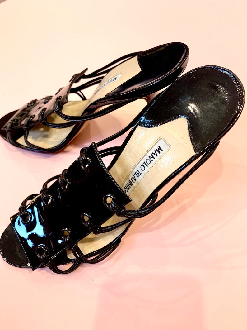 May include: A pair of black patent leather heeled sandals with a lace-up design. The sandals have a thin strap across the toe and a thick strap around the ankle. The brand name "MANOLO BLAHNIK" is visible on the insole.