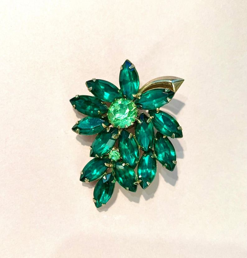 Pode incluir: Um broche verde e dourado com um grupo de strass em forma de marquise. O broche tem a forma de uma flor com um strass verde central.