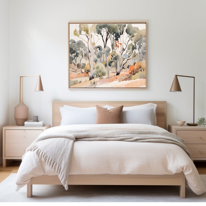 May include: A white bed with a beige blanket and a brown throw pillow. The bed is in a room with a white wall and a painting of trees above the bed.