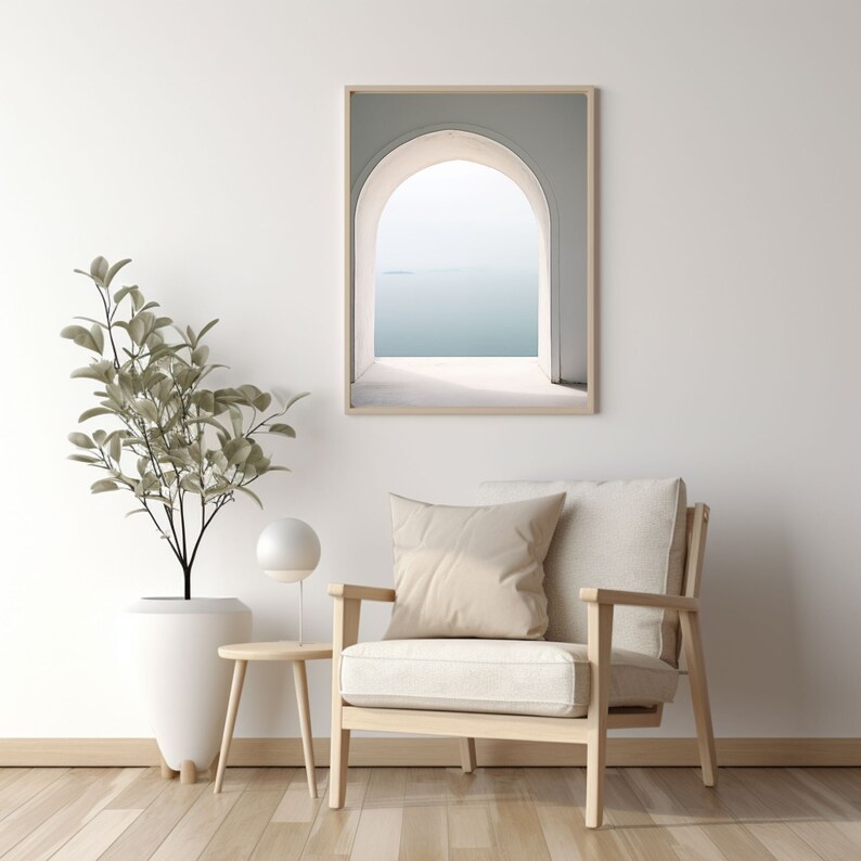 May include: A white armchair with a beige cushion sits in front of a white wall with a framed print of an archway overlooking a body of water.