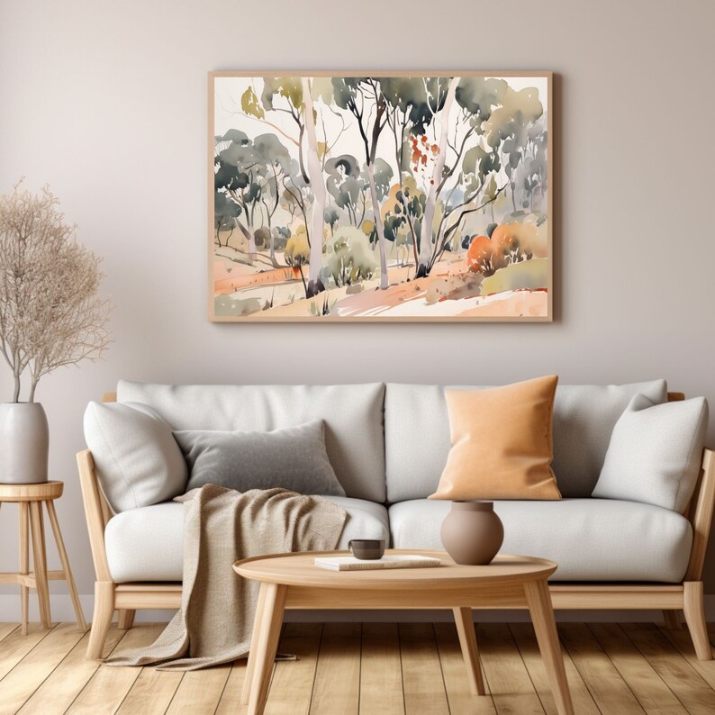 May include: A watercolor painting of a forest scene with tall trees and a light brown background. The painting is framed in a light wood frame and is hanging on a white wall above a white sofa with pillows and a throw blanket.